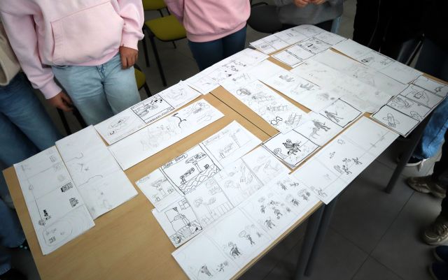 Na zdjęciu znajdują się dwa złączone stoły, na których leżą stworzone przez uczniów komiksy. Dookoła stolików stoją uczniowie.