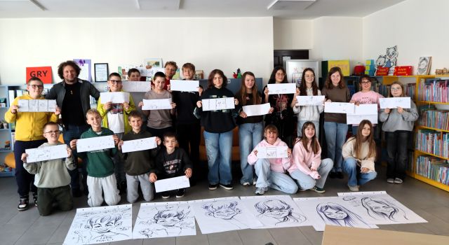 Na zdjęciu znajduje się 20 uczniów, którzy stoją do zdjęcia ze swoimi komiksami, wraz z nimi stoi mężczyzna, przed młodzieżą znajduje się 6 karykatur. W tle znajdują się półki z książkami.