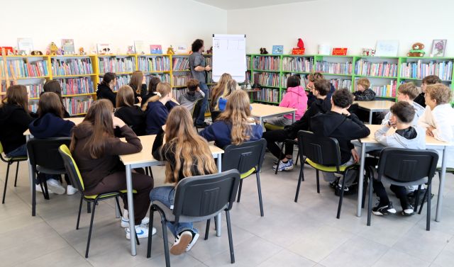 Na zdjęciu znajduje się 24 uczniów przy 5 stolikach, przed nimi stoi mężczyzna i jedną ręką trzyma stojący obok flipart. W tle znajdują się półki z książkami.