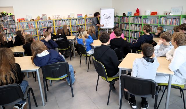 Na zdjęciu znajdują się uczniowie przy stolikach, przed nimi stoi mężczyzna i rysuje na fliparcie karykaturę chłopca. W tle znajdują się półki z książkami.