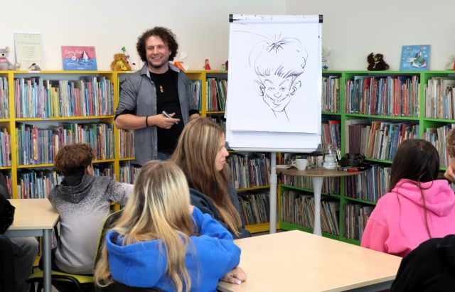 Na zdjęciu znajdują się dwa stoliki, przy nich 4 nastolatków, przed nimi stoi mężczyzna, a obok niego flipart, na którym namalowana jest karykatura chłopaka, obok stoi stolik z wodą. W tle znajdują się półki z książkami.
