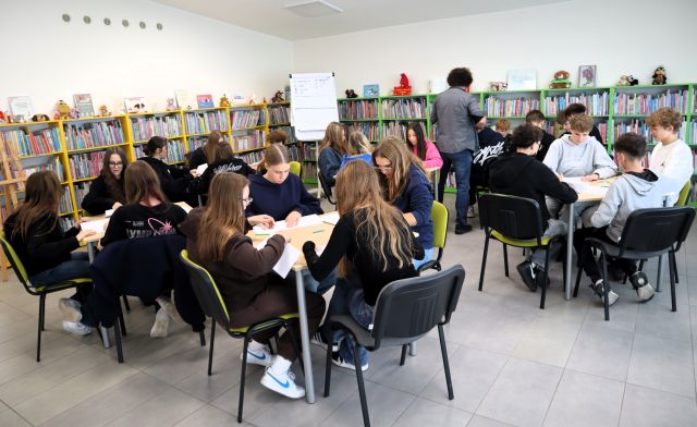 Na zdjęciu znajduje się 24 uczniów przy 5 stolikach i rysują na kartkach. Przy jednym ze stolików stoi mężczyzna. W tle znajduje się flipart oraz półki z książkami.