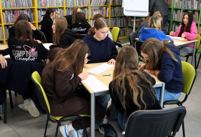 Na zdjęciu w centrum znajdują się 4 dziewczyny przy stoliku i rysują na kartkach swój komiks. W tle znajdują się inni uczestnicy zajęć przy stolikach oraz półki z książkami.