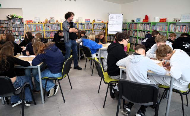 Na zdjęciu znajduje się 5 stolików, a przy nich 24 uczniów, którzy rysują na kartkach swój komiks, między stolikami stoi mężczyzna. W tle znajduje się flipart oraz półki z książkami.
