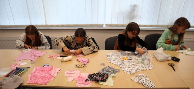 Na zdjęciu znajdują się cztery dziewczynki, które odrysowują wzór na materiałach przy złączonych stołach, na których leżą kolorowe materiały. W tle znajdują się okna.