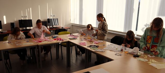 Na zdjęciu znajduje się 6 dziewczynek przy złączonych stołach w literę C i wycinają z kolorowych materiałów wcześniej odrysowany wzór. Na stole znajdują się nożyczki, nici i kolorowe materiały. Warsztaty odbywają się w sali komputerowej.