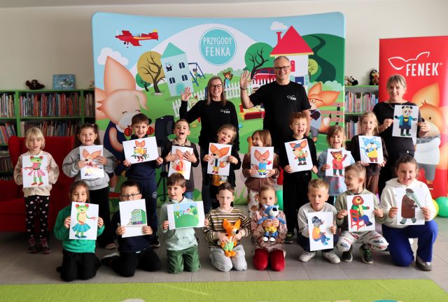 Na zdjęciu znajduje się 17 przedszkolaków oraz dwie kobiety i mężczyzna, dzieci oraz jedna z kobiet trzymają kartki z postaciami z książek o przygodach fenka, dwoje dzieci trzyma małe maskotki. Mężczyzna i kobieta z tyłu podnoszą rękę do góry. Za nimi znajduje się baner z ilustracja bohaterów na placu zabaw oraz czerwony baner po prawej stronie. W tle znajdują się półki z książkami.