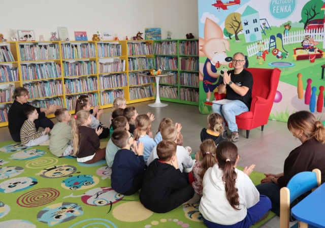 Na zdjęciu znajdują się przedszkolaki na dywanie oraz jedna kobieta i klaszczą wraz z mężczyzną, który siedzi naprzeciwko nich na fotelu, za mężczyzną znajduje się baner z bohaterami książki, którzy bawią się na placu. W tle znajdują się półki z książkami.