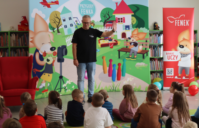 Na zdjęciu znajdują się dzieci na dywanie, przed nimi stoi mężczyzna i im macha, za mężczyzną stoi baner z ilustracją z książek o Fenku, po prawo czerwony baner z książkowym lisem Fenkiem, a po lewo czerwony fotel. W tle znajdują się półki z książkami.