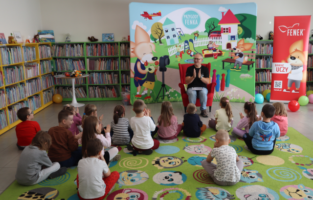 Na zdjęciu znajdują się dzieci na dywanie, przed nimi siedzi mężczyzna na foteli i wszyscy klaszczą. Za mężczyzną znajduje się baner z postaciami z książki na placu zabaw, po prawej stronie znajduje się czerwony baner z lisem z książki. W tle znajdują się półki z książkami.