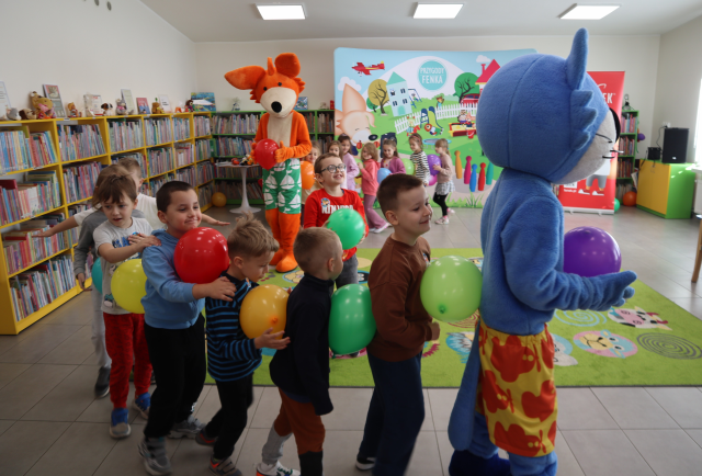 Na zdjęciu znajdują się dzieci, które robią pociąg za lisem i niebieskim kotem, a pomiędzy sobą na poziomie brzucha mają balony. W tle znajdują się półki z książkami oraz baner z postaciami z bajki na placu zabaw.