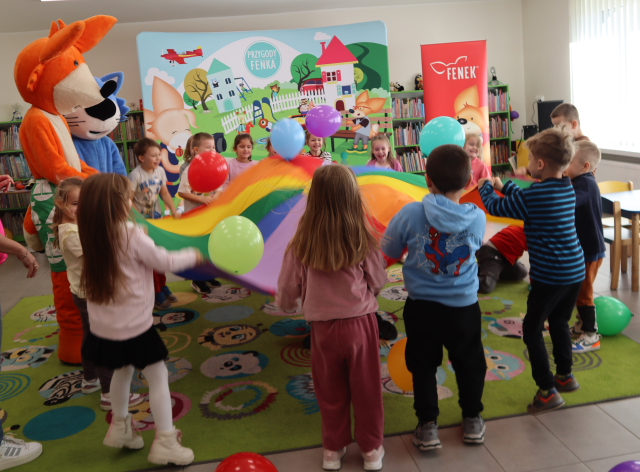 Na zdjęciu znajdują się dzieci trzymające kolorową chustę wraz z maskotka niebieskiego kota i lisa i podrzucają balony. W tle znajdują się półki z książkami oraz baner z postaciami książkowymi na placu.
