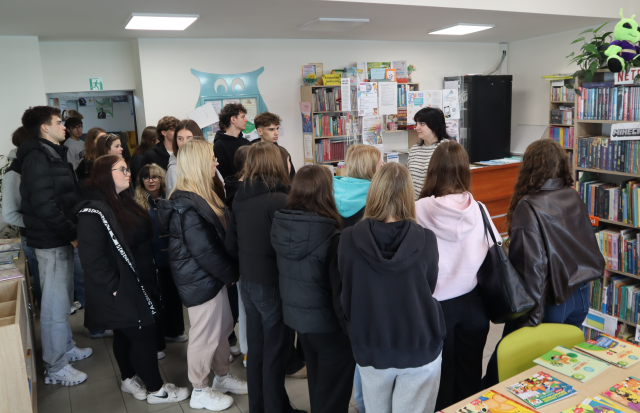 Na zdjęciu znajduje się młodzież w sali bibliotecznej, po prawej stronie przy półce z książkami stoi kobieta. W tle znajduje się biurko oraz drzwi.