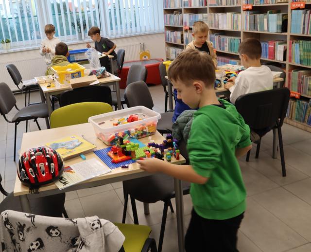 Na zdjęciu znajduje się 7 chłopców przy 4 stolikach, przy których układają klocki LEGO, które znajduje się w pudełkach na stolikach. W tle znajdują się półki z książkami oraz okna.