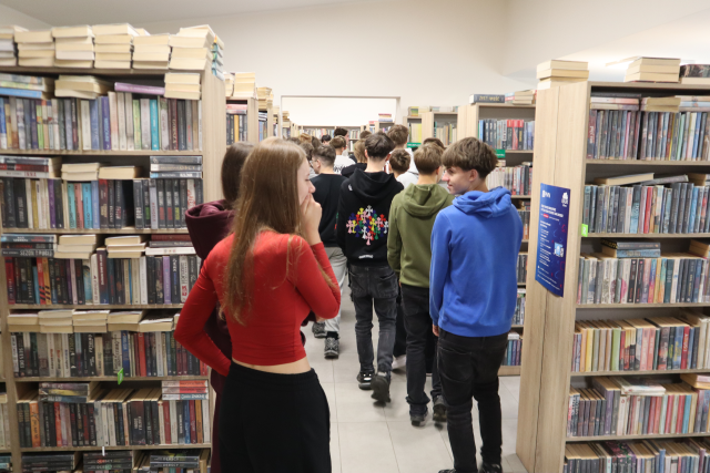 Na zdjęciu znajduje się grupka młodzieży między półkami z książkami w bibliotece.