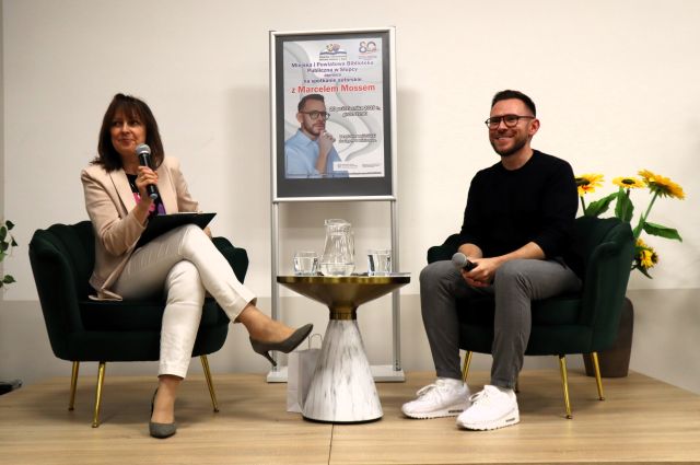 Na zdjęciu znajduje się mała scena, na której siedzi kobieta i mężczyzna na fotelach, oboje trzymają mikrofony, pomiędzy nimi stoi stolik z wodą, a za nim plakat promujący spotkanie. Po bokach sceny znajdują się sztuczne kwiaty.