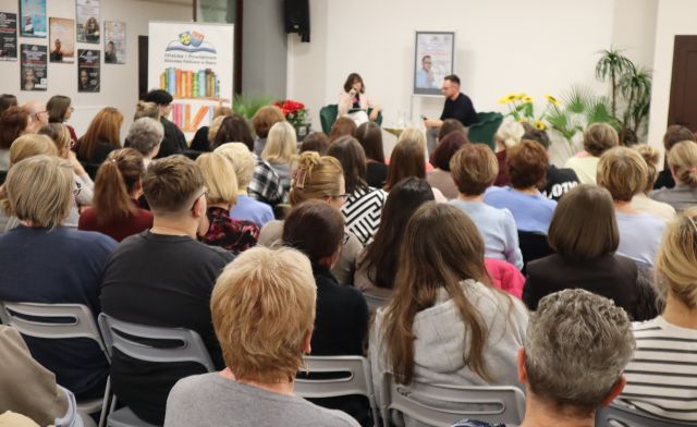 Na zdjęciu znajduje się widownia na holu biblioteki, przed nimi siedzi kobieta i mężczyzna na scenie, za nimi znajduje się plakat promujący spotkanie. W tle znajdują się kwiaty, baner z logiem biblioteki i książkami, drzwi.