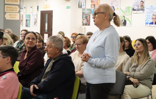 Na zdjęciu znajduje się widownia na holu biblioteki, jedna z kobiet stoi. W tle znajdują się drzwi, plakaty ścianach. Zdjęcie zostało zrobione od boku.
