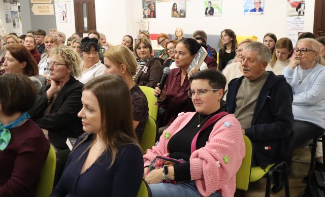 Na zdjęciu znajduje się widownia na holu biblioteki, jedna z kobiet pokazuje książkę i trzyma mikrofon przy ustach. W tle znajdują się plakaty na ścianach, drzwi.