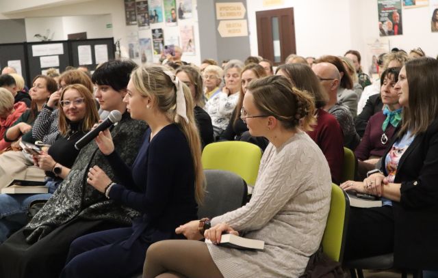 Na zdjęciu znajduje się widownia na holu biblioteki, jedna z kobiet z pierwszego rzędu mówi przez mikrofon. W tle znajdują się drzwi, czarna tablica z wystawką, plakaty na ścianach.