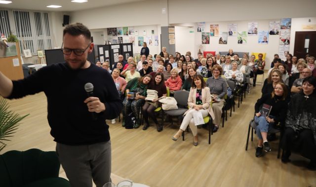 Na zdjęciu znajduje sie mężczyzna, który stoi na scenie i robi sobie zdjęcie z widownią , która siedzi na holu biblioteki. W tle znajdują się plakaty na ścianach.
