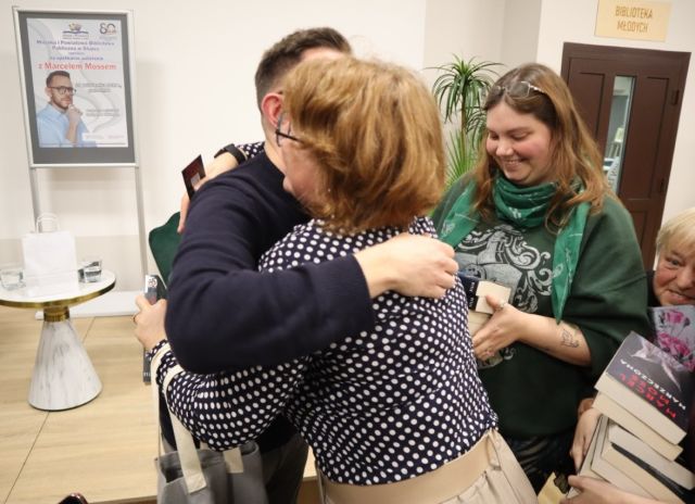 Na zdjęciu znajduje się mężczyzna oraz kobieta i się przytulają, po prawej stronie stoją dwie kobiety z książkami, po lewo znajduje się scena ze stolikiem i plakatem promującym spotkanie.