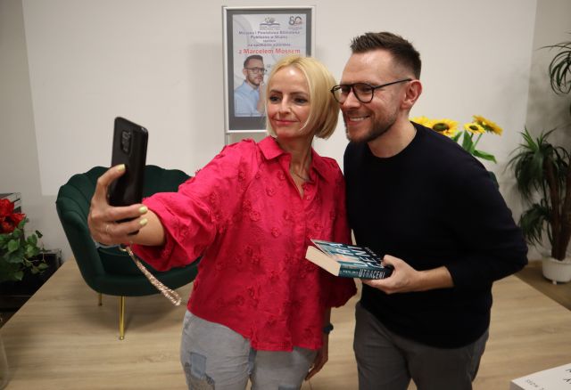 Na zdjęciu znajduje się mężczyzna oraz kobieta, mężczyzna trzyma książkę, a kobieta telefon i robi im wspólne zdjęcie. W tle znajduje się scena, na której stoją fotele i plakat promujący spotkanie.