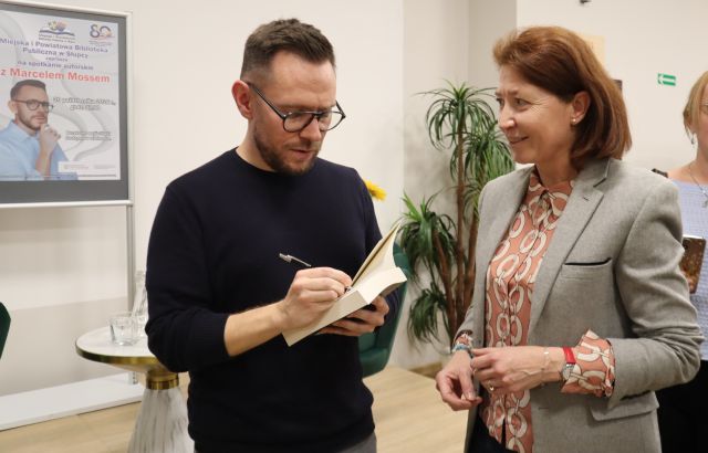 Na zdjęciu znajduje się mężczyzna, który podpisuje stojącej obok kobiecie książkę. W tle znajduje się plakat promujący spotkanie oraz kwiaty.