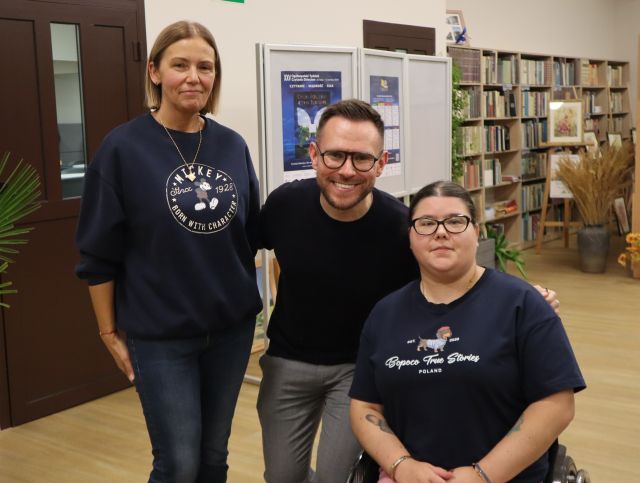 Na zdjęciu znajdują się dwie kobiety oraz mężczyzna i uśmiechają się do wspólnego zdjęcia. W tle znajdują się drzwi, plakaty oraz półki z książkami.