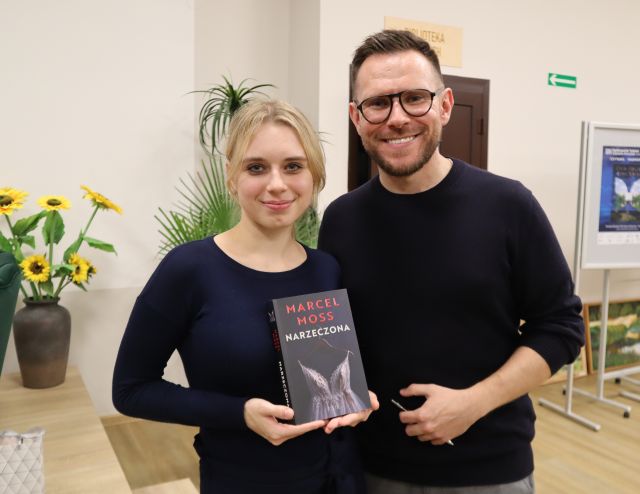 Na zdjęciu znajduje się mężczyzna i kobieta z książką, którzy stoją do wspólnego zdjęcia. W tle znajdują się kwiaty, drzwi, plakaty.