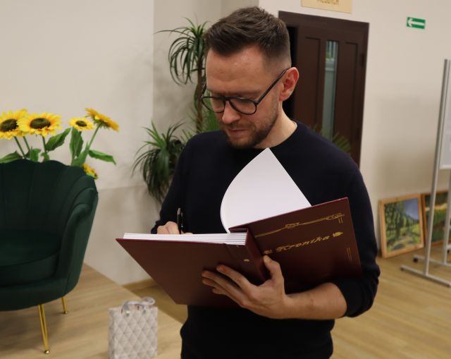 Na zdjęciu znajduje sie mężczyzna, który stoi i wpisuje się do kroniki. W tle znajdują się kwiaty, drzwi i fotele na scenie po lewej stronie.