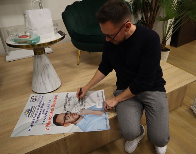 Na zdjęciu znajduje się mężczyzna siedzący na scenie i podpisujący plakat, obok niego stoi stolik i fotele.