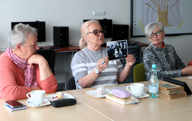 Na zdjęciu znajdują się 3 kobiety przy złączonych stolikach, kobieta po środku pokazuje czarno-białą fotografię. Na stolikach leżą filiżanki, pokrowce na okulary, woda, stare zdjęcia, książki. Warsztaty odbywają się w sali komputerowej.
