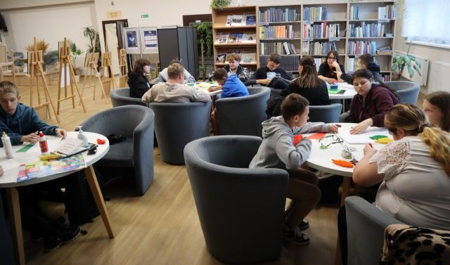Na zdjęciu znajduje się młodzież przy 4 stolikach w czytelni biblioteki i wycinają z kolorowych kartek. W tle znajdują się półki z książkami, po prawo okna, a po lewo czarna tablica, plakaty i drzwi.