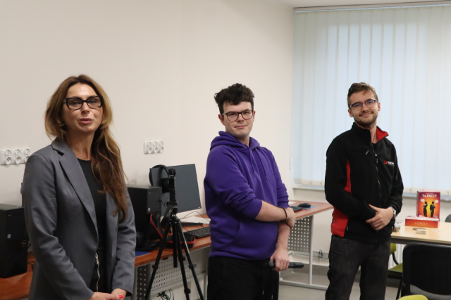 Na zdjęciu znajduje się jedna kobieta oraz dwóch mężczyzn stojących obok siebie, pomiędzy kobietą a mężczyzną stoi mała kamerka na statywie. W tle znajduje się stolik z grą Tajniacy. Warsztaty odbywają się w sali komputerowej.