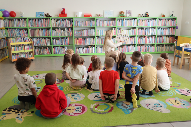 Na zdjęciu znajdują się dzieci na dywanie, a przed nimi siedzi kobieta i pokazuje dzieciom ilustracje z książki. W tle znajdują się półki z książkami.