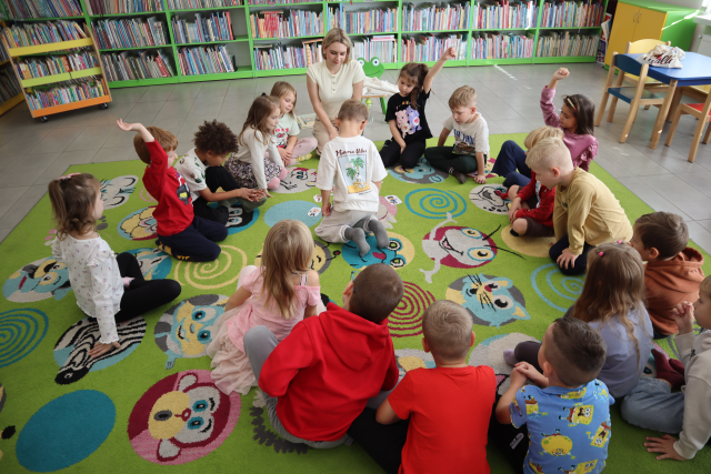 Na zdjęciu znajdują się dzieci wraz z kobietą na dywanie, część dzieci ma podniesioną do góry rękę. W tle znajdują się półki z książkami.