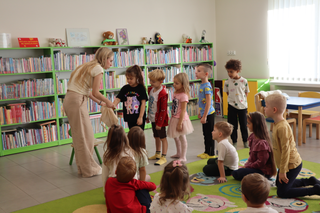 Na zdjęciu znajdują się dzieci na dywanie, przed nimi stoi kobieta i pochyla się z torbą z której po kolei 5 dzieci stojąca w kolejce losuje przedmioty. W tle znajdują się półki z książkami i mały stolik.