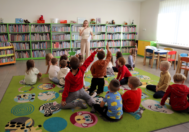 Na zdjęciu znajdują się dzieci na dywanie, część z nich podnosi do góry rękę, przed nimi stoi kobieta i w ręce trzyma kartkę. W tle znajdują się półki z książkami, małe stoliczki po prawo.