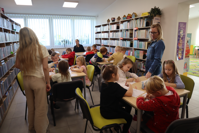 Na zdjęciu znajdują się przedszkolaki przy 6 stolikach i rysują na kartkach, między stolikami znajdują się 3 kobiety. W tle znajdują się okna, a po bokach półki z książkami.