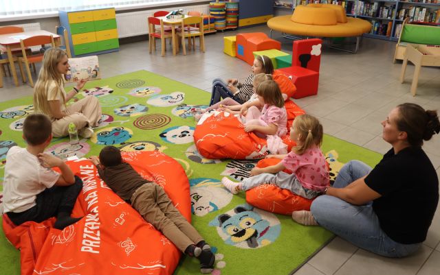 Na zdjęciu znajduje się 6 dzieci na dywanie na pufach wraz z jedną kobietą, przed nimi siedzi kobieta i pokazuje im ilustracje z książki. W tle znajdują się małe stoliki, pufy ułożone w pociąg, okrągła kanapa oraz półki z grami.
