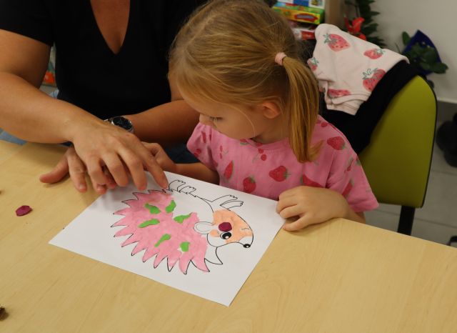 Na zdjęciu znajduje się dziewczynka, która ozdabia plasteliną wydrukowanego na kartce jeżyka, obok niej siedzi mama.