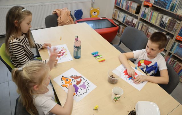Na zdjęciu znajduje się 3 dzieci przy złączonych stolikach i ozdabiają wydrukowanego na kartce języka plasteliną, na stoliku znajdują się dwa plastikowe pojemniki, plastelina i bidon. W tle znajdują się półki z książkami oraz stolik interaktywny.