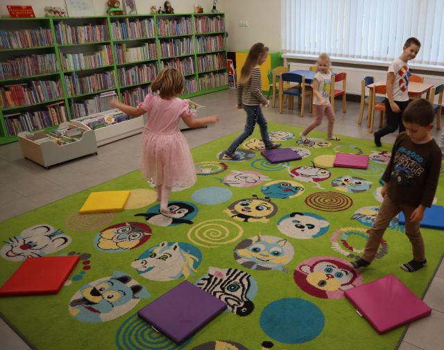 Na zdjęciu znajduje się 5 dzieci, chodząca po dywanie i omijająca leżące na nim pufy. W tle znajdują się półki z książkami, stoliki z krzesełkami oraz okna.