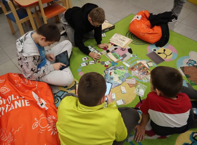 Na zdjęciu znajduje się 4 chłopców, którzy siedzą na dywanie, obok nich leżą czerwone pufy i grają w grę planszową, jeden z chłopców trzyma tablet.