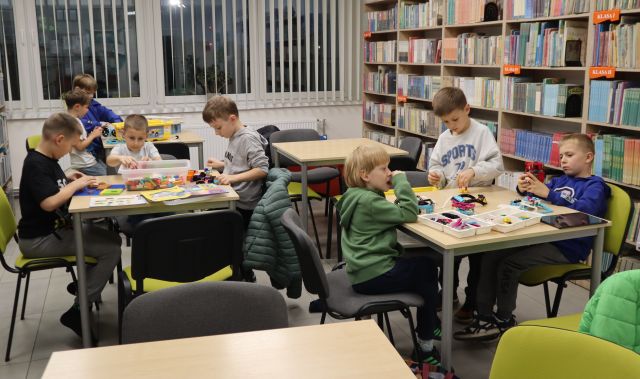 Na zdjęciu znajduje się 8 chłopców przy 3 stolikach, dwa stoły obok są puste. Chłopcy układają zestawy LEGO. W tle znajdują się półki z książkami i okna.