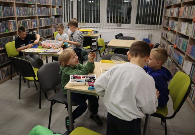 Na zdjęciu znajduje się 6 chłopców przy 2 stolikach, dwa stoły są puste i chłopcy układają klocki LEGO. W tle znajdują się półki z książkami i okna.