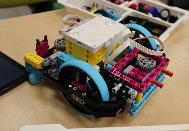 Na zdjęciu znajduje się samochód zbudowany z klocków LEGO, który leży na stoliku, obok niego leży tablet oraz plastikowe pudełko z klockami.