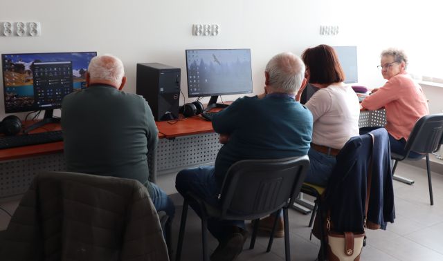 Na zdjęciu znajdują się 2 kobiety i 2 mężczyzn przy biurkach z 3 komputerami. Na krzesłach wiszą kurtki. Warsztaty odbywają się w sali komputerowej.