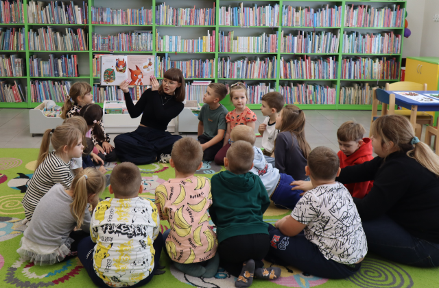 Na zdjęciu znajduje się 16 przedszkolaków, którzy siedzą w okręgu, po prawej stronie wraz z nimi siedzi kobieta oraz między nimi, na przeciwko i pokazuje im ilustracje z książki z wiewiórką. W tle znajdują się półki z książkami.